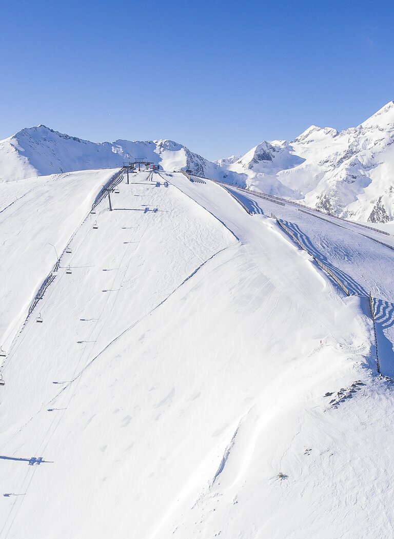 Aerial view of a freshly groomed ski slope with chairlift on a snowy alpine ridge under clear blue sky. | © Galsterbergalm Bahnen GmbH & Co KG 