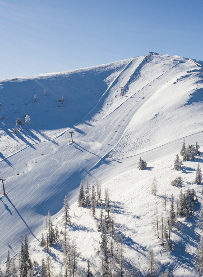 Wide groomed ski slope crosses a snow-covered mountain ridge with ski lifts and scattered trees. | © Galsterbergalm Bahnen GmbH & Co KG 