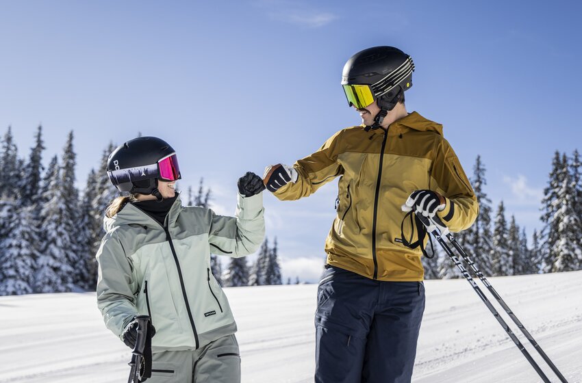 Zwei Skifahrer mit Helmen und Skistöcken begrüßen sich auf einer verschneiten Piste vor winterlichen Nadelbäumen. | © Mirja Geh
