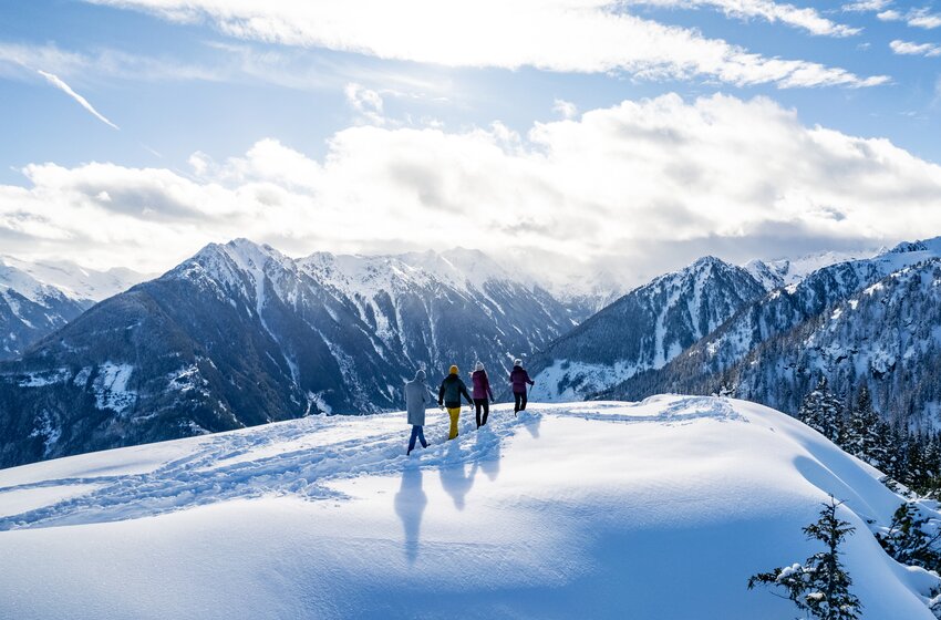 Vier Personen wandern durch frischen Pulverschnee mit Blick auf verschneite Berggipfel und winterliche Alpenlandschaft. | © Winterwanderbilder (c) Gerald Oberreiter