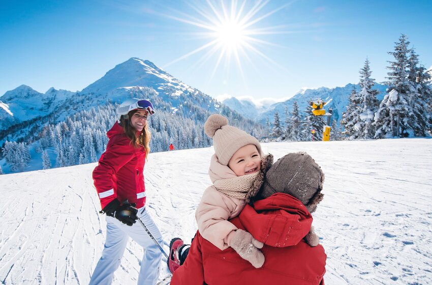Frau mit Skiern und Kind in Winterkleidung auf Skipiste, strahlende Sonne und verschneite Berge im Hintergrund. | © Ski amadé GmbH